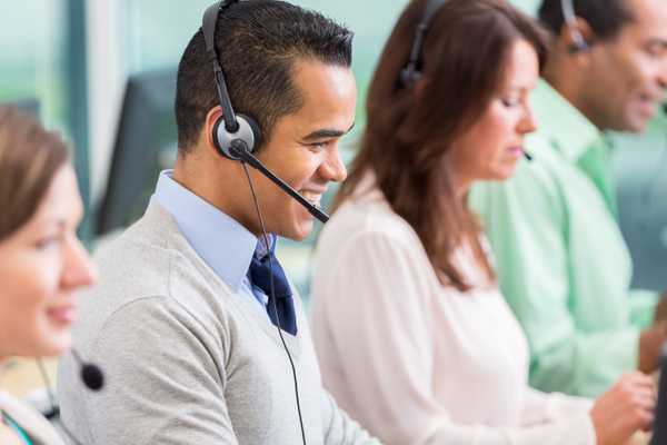 Cheerful mid adult Hispanic male IT support person smiles while talking with customer on the phone. He is wearing a headset. His colleagues are working in the foreground and the background.